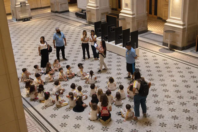 Um grupo de crianças está sentado no chão da Sala São Paulo em roda enquanto os educadores contam curiosidades sobre o local.
