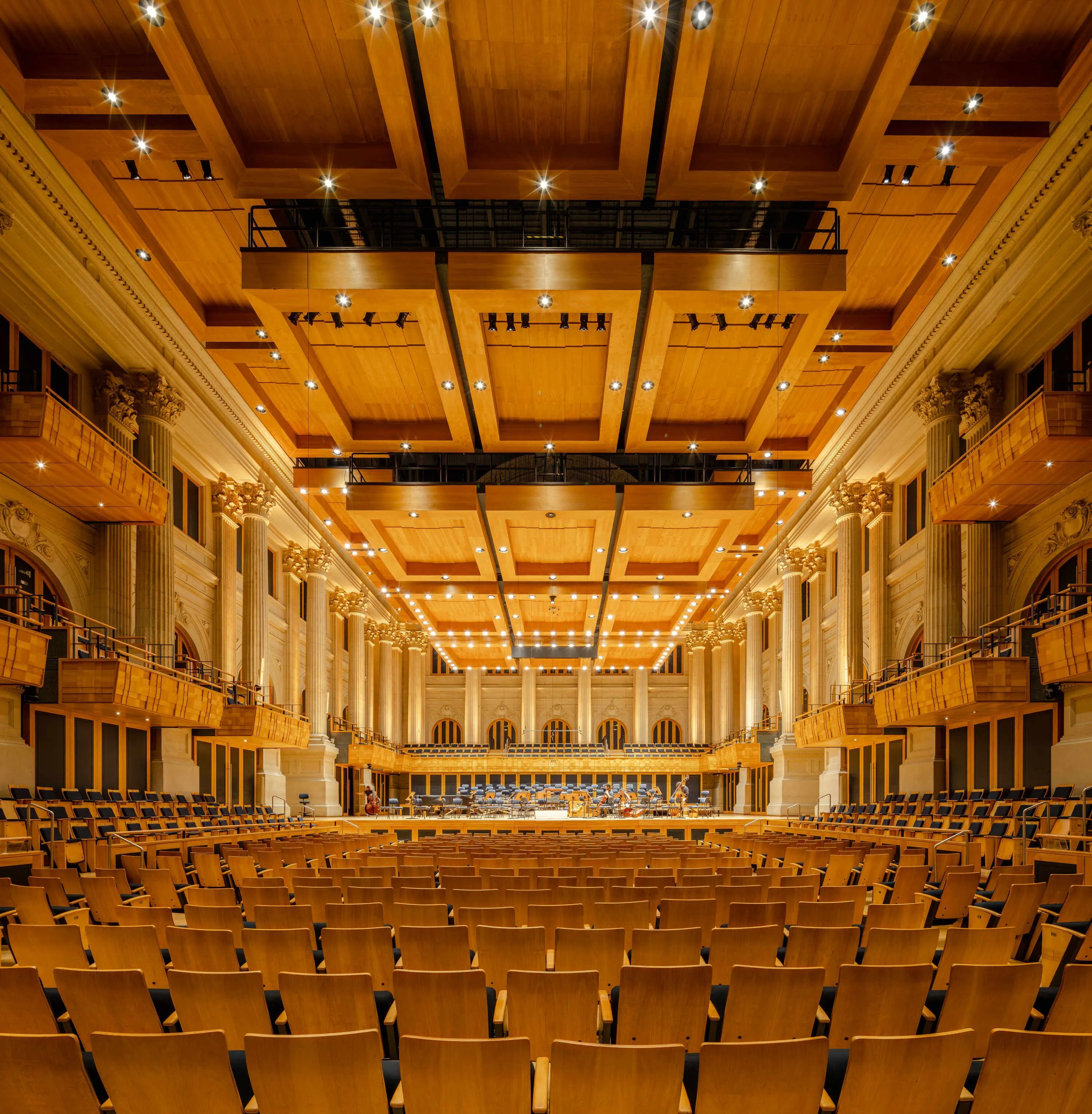 A imagem mostra a vista da Sala São Paulo, com o teto móvel, colunas iluminadas e cadeiras de madeira amarela com estofado azul.