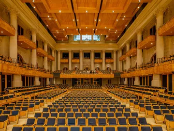 Sala de concertos com pessoas na plateia e no palco. As colunas estão iluminadas de azul e o palco com uma luz quente.
